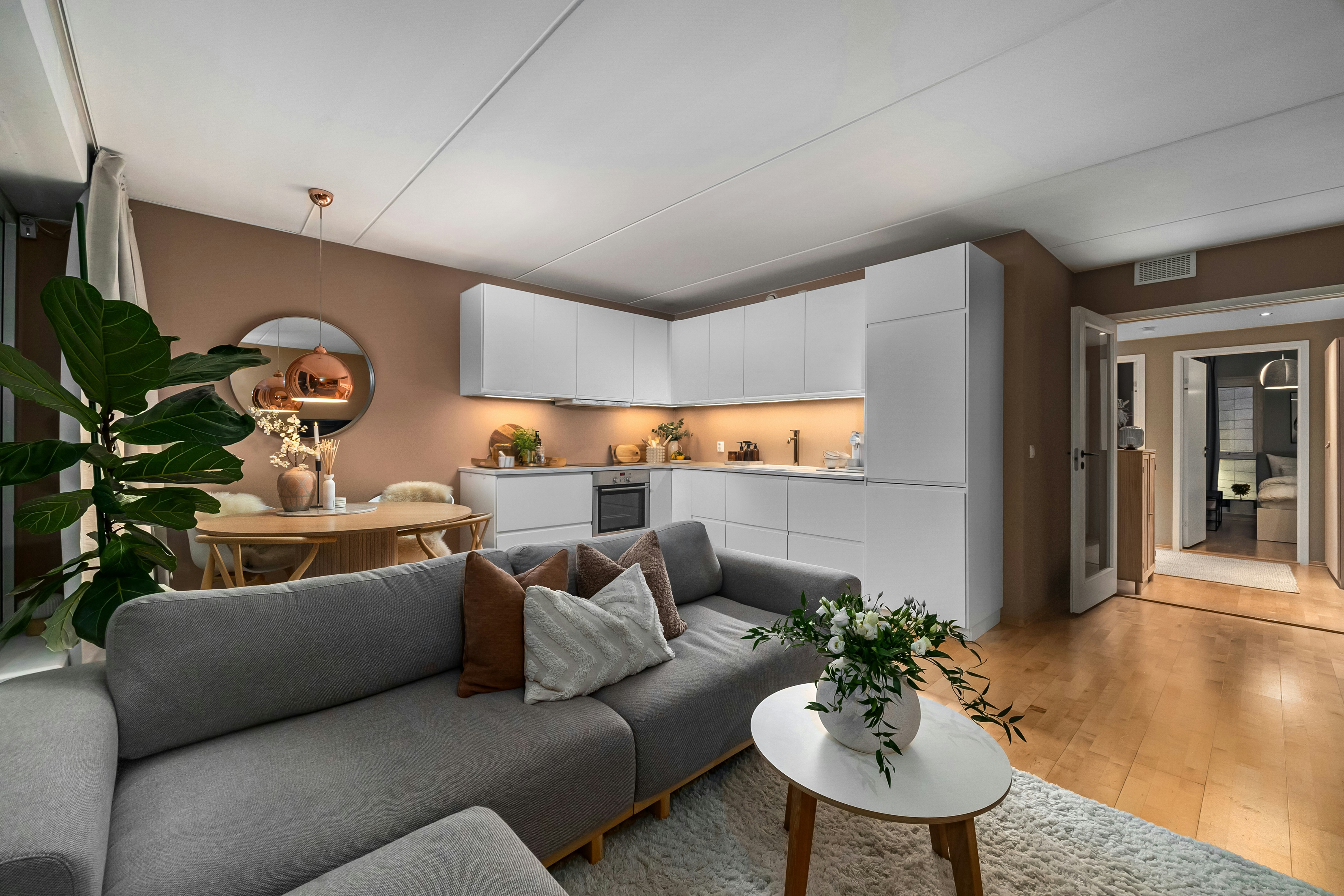 This image depicts a kitchen and living space with light brown walls. A grey sofa, wooden furniture and white cabinetry compliment the brown walls.