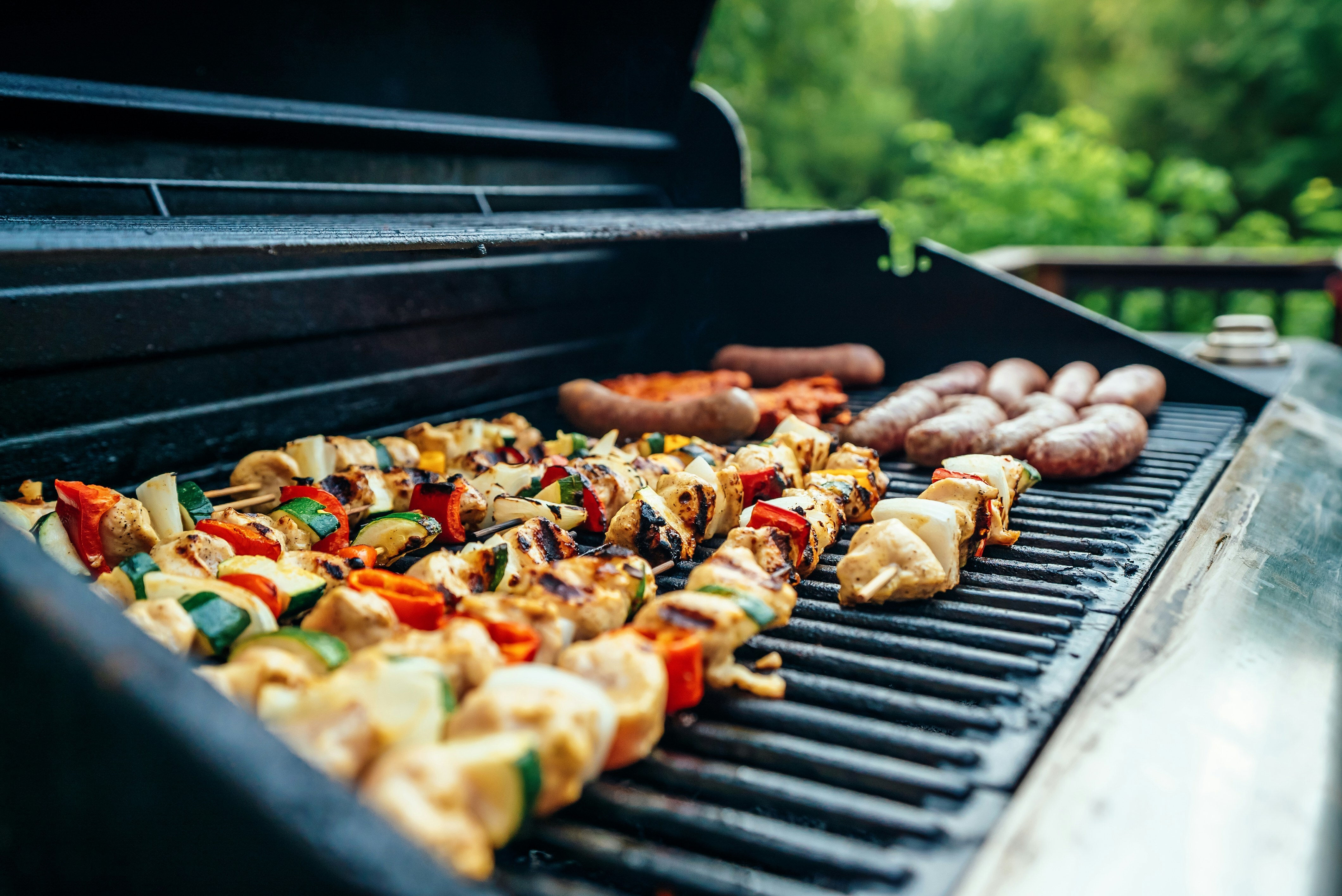 A grill has kabobs and sausages on it for an outdoor cookout.